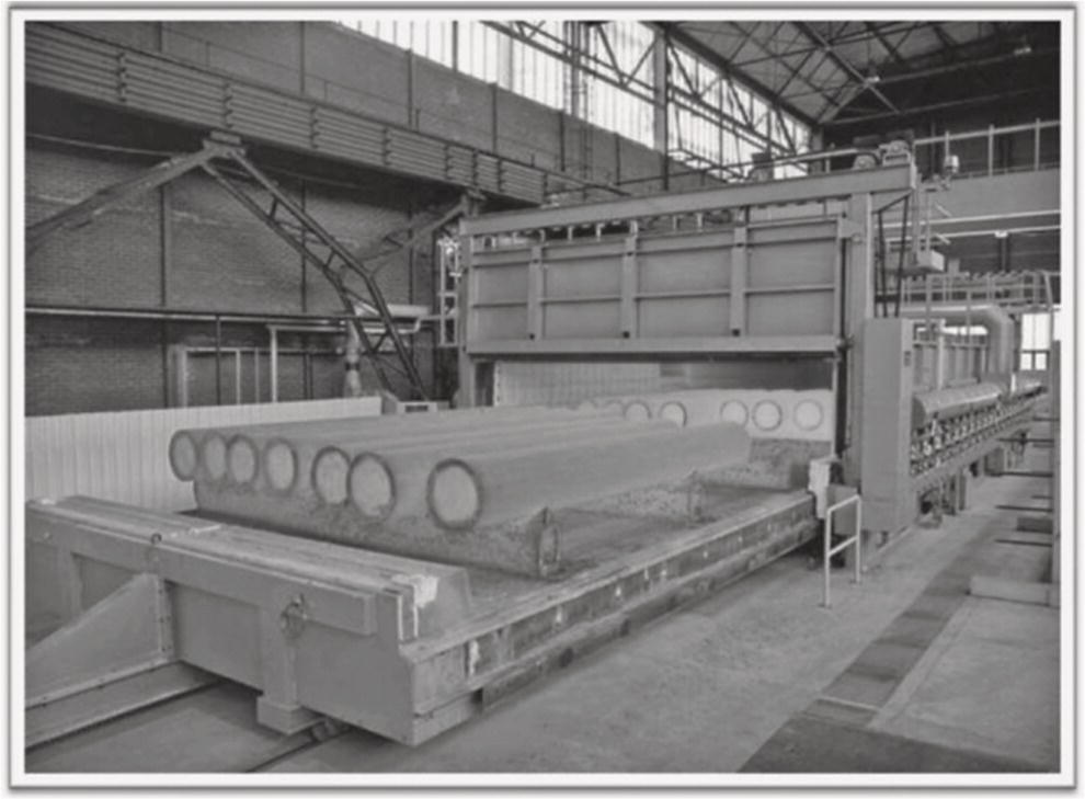 Image of an industrial factory interior with large machinery. The main focus is on a large furnace-like structure containing long cylindrical objects. Overhead, metal beams and a high ceiling are visible.