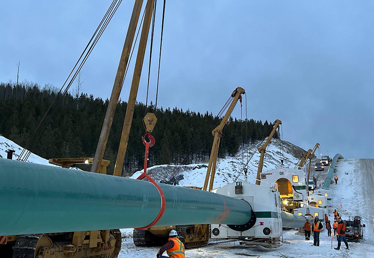 A photograph of a construction worker installing a large pipeline in a snow-covered area. Several cranes are positioned along the pipeline, holding it in place. Workers in safety gear are scattered throughout the scene.