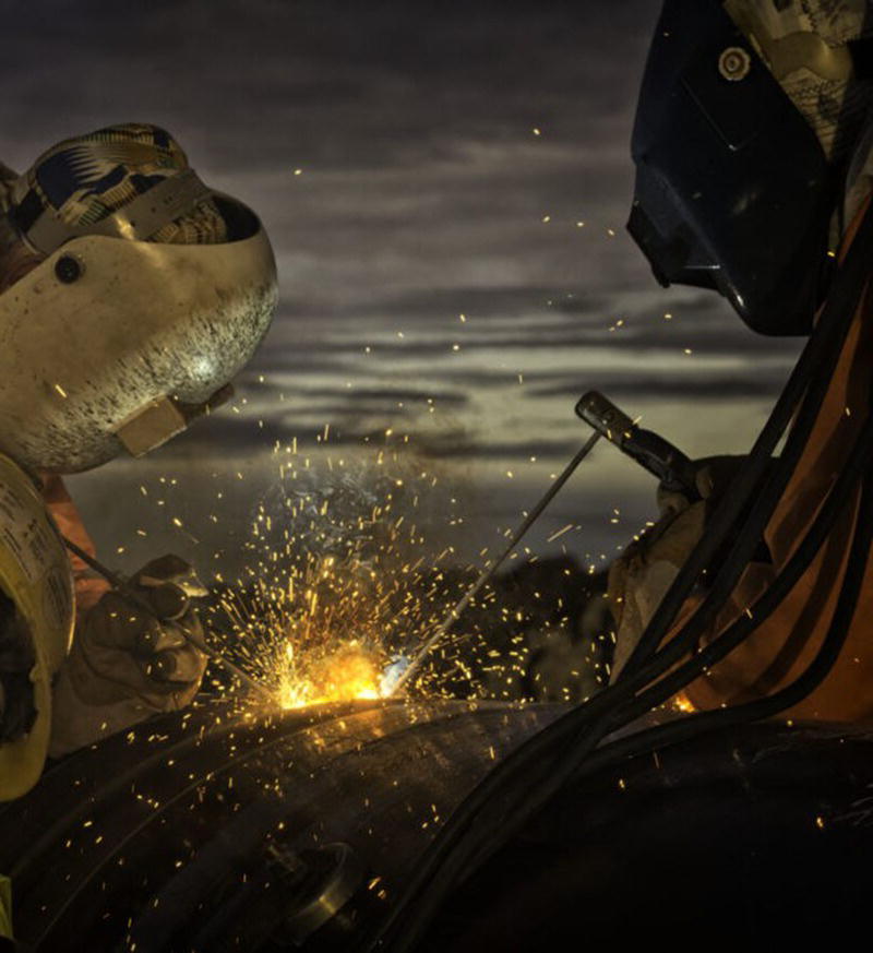 A photograph shows two welders wearing protective helmets and suits working on a large metal pipe, creating a bright burst of sparks.