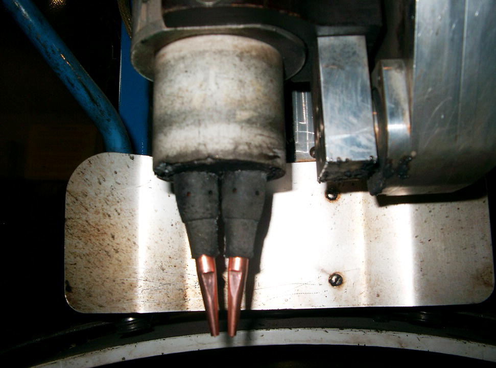 A close-up image of industrial machinery, featuring two conical metal nozzles attached to a cylindrical component. The machine’s surfaces show signs of wear with dark smudges and scratches.