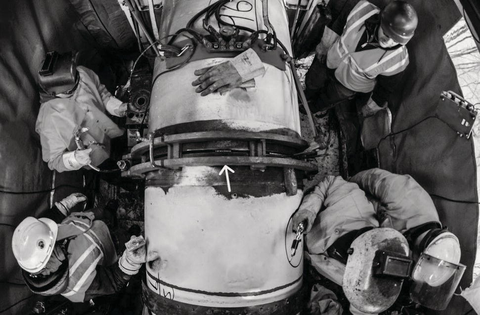 Overhead view of five workers wearing safety gear and helmets, gathered around a large horizontal pipe. One worker uses a tool on the pipe, while others observe or assist. An arrow is marked on the pipe. Protective coverings surround the work area.