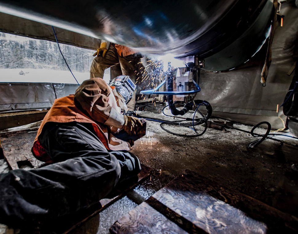A photograph of a welder wearing protective gear lies on the ground, working on a large metal pipe. Sparks fly from the welding torch. The background has equipment.
