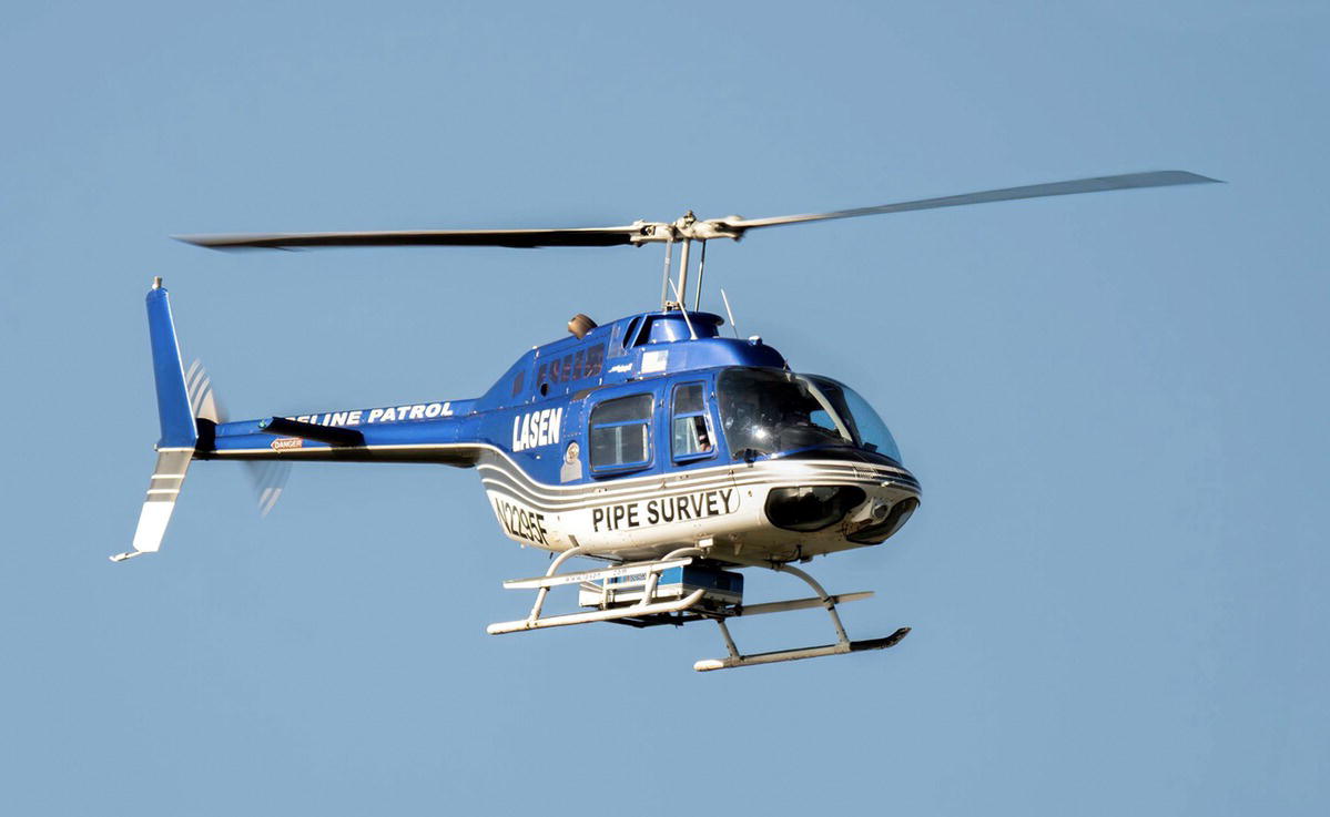 A photograph of a helicopter marked with lasen, pipeline patrol, and pipe survey is in flight against a clear sky. The helicopter has visible landing skids. Its rotor blades are in motion.