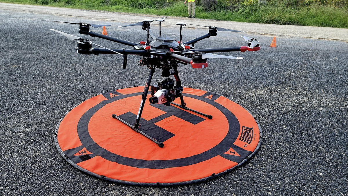 A photograph of a drone with six propellers is positioned on a circular launch pad marked with a large H. The drone has a mounted camera and stands on a tripod-like landing gear.