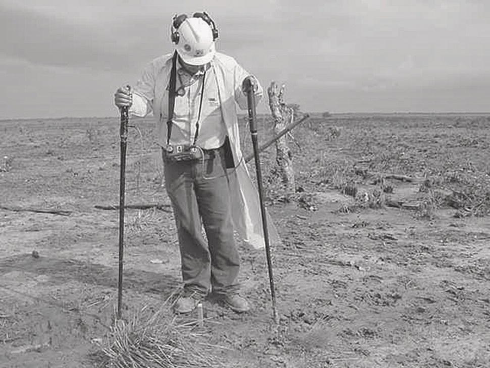 A person stands in a barren landscape covered with uneven, cracked earth. They wear a helmet, headphones, and a light-coloured shirt, complemented by dark pants and sturdy boots. Their hands grip two long poles that are thrust into the ground, while a camera hangs around their neck. Sparse vegetation struggles to grow in the dry soil, hinting at a challenging environment. The sky above is cloudy, creating a sombre atmosphere, and remnants of stumps or trees are visible in the background, suggesting a previously forested area.