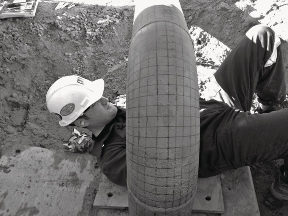 A construction worker in a hard hat lies on his back in a dug-out area, adjusting or inspecting a large pipe. The pipe, prominent in the foreground, has a textured surface with a grid-like pattern. The worker’s torso is visible, dressed in a dark outfit, while one arm extends towards the pipe, possibly handling tools or equipment. The surrounding ground is uneven, indicating an excavation site, and there are scattered items in the background, suggesting an active work environment. Sunlight casts shadows, enhancing the scene’s depth.