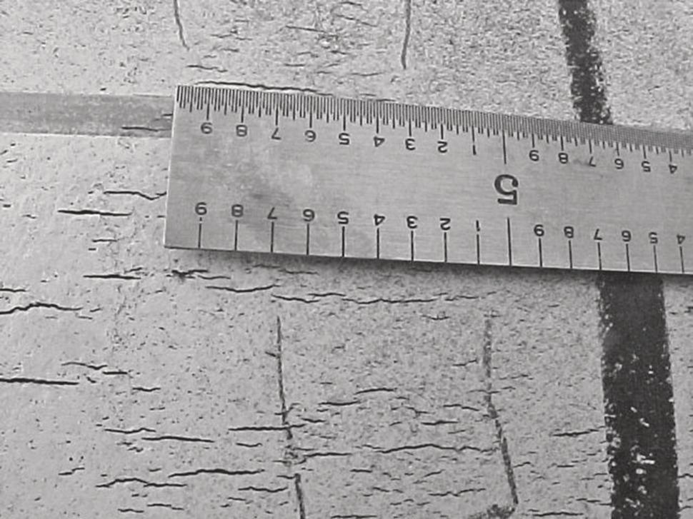 A ruler rests on a textured surface marked with lines. The ruler, measuring in centimetres, indicates a length of five centimetres at the beginning. The underlying surface appears worn and has a cracked appearance, with a prominent dark line running vertically. Smaller scratches or marks are also visible, adding to the aged look of the material. The composition comprises a stark contrast between the metallic surface of the ruler and the unevenness of the ground below it, highlighting the measurement in a practical context.