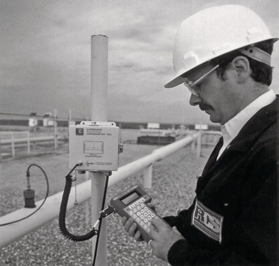 A worker, wearing a hard hat and safety glasses, stands outdoors in an industrial setting. He examines a handheld device in his left hand while his right hand interacts with a panel mounted on a vertical pipe. This panel appears to have a digital display and various buttons. The background features a network of pipes and equipment, indicative of a manufacturing or processing facility. The scene conveys a sense of maintenance or monitoring activity, emphasizing the technological aspect of industrial operations.