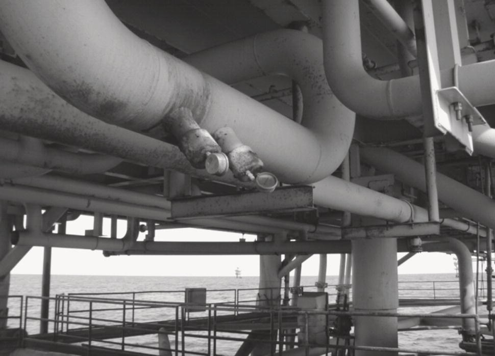 A network of industrial pipes runs beneath a structure, intersecting at various angles and creating a complex arrangement. The pipes appear weathered, indicating exposure to harsh environmental conditions. The background reveals an expansive ocean, with a distant oil platform visible on the horizon. Structural supports maintain the framework, with railings suggesting accessibility for maintenance or operations. The scene conveys a sense of industry and engineering, highlighting the infrastructure essential for offshore activities.