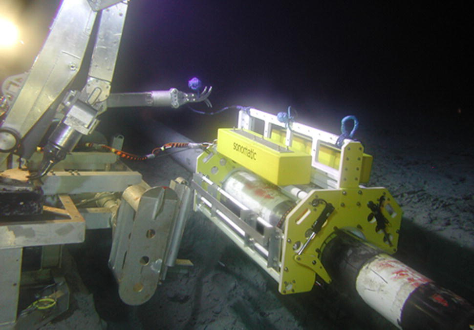A complex underwater scene features a robotic arm positioned near a cylindrical device resting on the seabed. The robotic arm extends towards the device, which is mounted on a larger frame with various attachments. The environment appears dark, illuminated by a light source, highlighting the textures of the sea floor and the machinery’s metallic surfaces. The device, possibly involved in underwater research or exploration, is equipped with sophisticated technology, indicating its use in deep-sea operations or scientific endeavours.