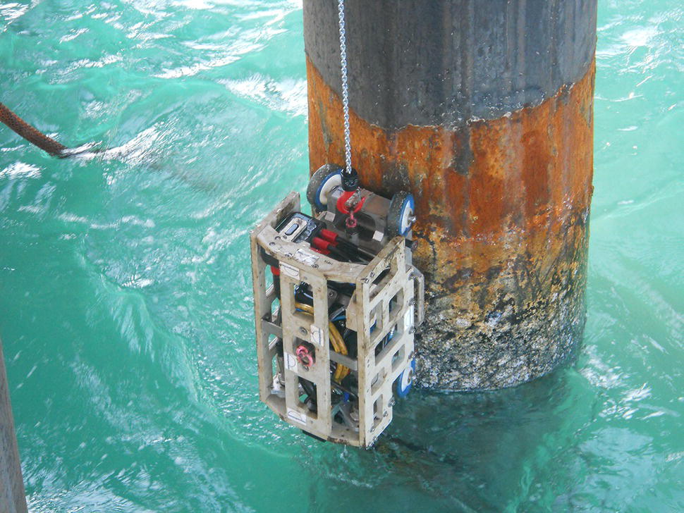 A structure hangs from a pier, submerged in water. This apparatus, resembling a cage, is attached to a vertical pole by a chain. The water’s surface reflects light, creating ripples around the base of the structure. The cage appears robust and functional, potentially designed for underwater operations. Its metallic components are visible, hinting at a mechanical purpose. The pier’s post displays signs of wear, with barnacles and discoloration from exposure to the elements.