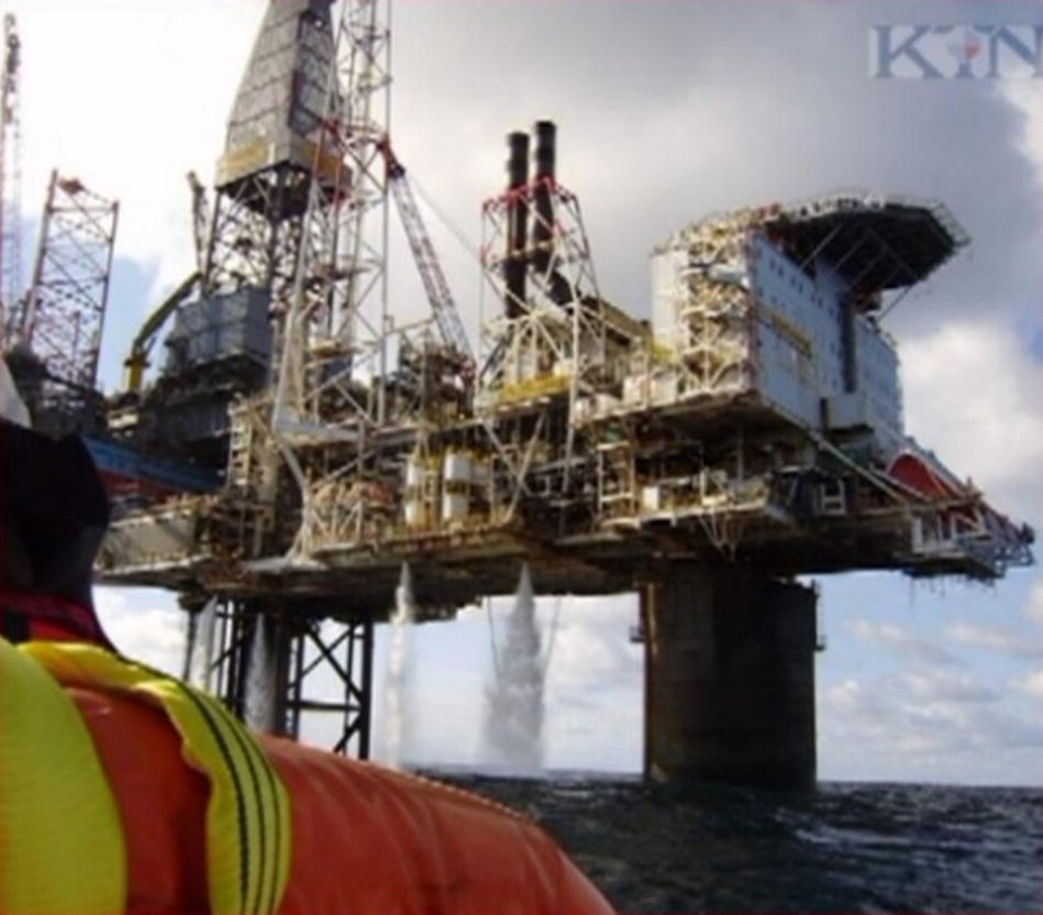 A large offshore oil rig stands prominently against a cloudy sky, supported by sturdy legs that extend into the ocean. Its structure features multiple platforms and compartments, with pipes and machinery visible. Two tall smokestacks rise from the rig, indicating active operations. In the foreground, a person in a life jacket looks towards the rig, creating a sense of scale and connection. The ocean appears calm, reflecting the complex engineering of the rig, which plays a crucial role in energy production.