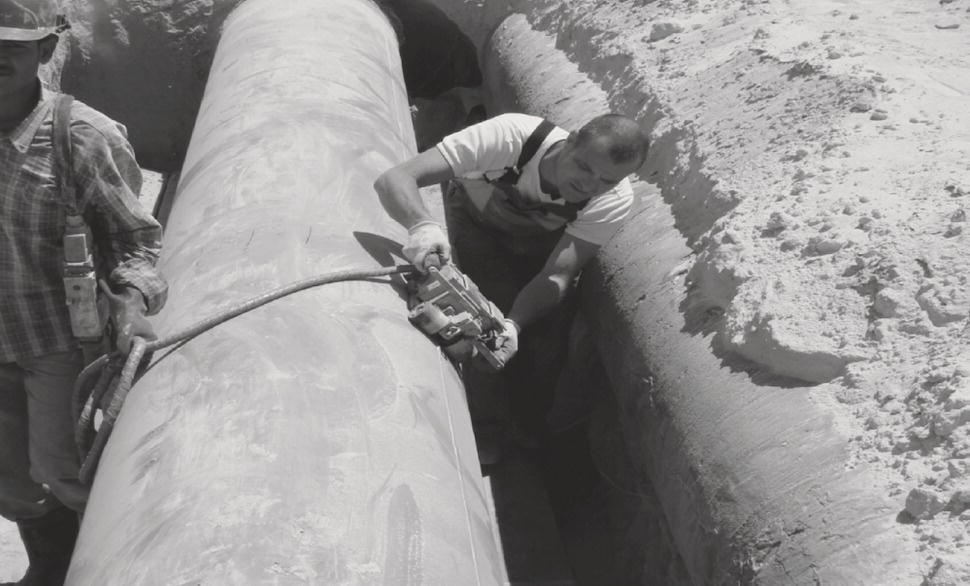 Two workers are engaged in a task at a construction site, focusing on a large cylindrical pipe positioned within a trench. One worker, equipped with a power tool, is actively cutting or modifying the pipe. He is dressed in a short-sleeve shirt and appears concentrated on the task. The second worker, standing nearby, wears a checked shirt and is observing the process. The surrounding area is sandy, indicating an excavation context, and the overall environment suggests a work zone dedicated to construction or infrastructure development.