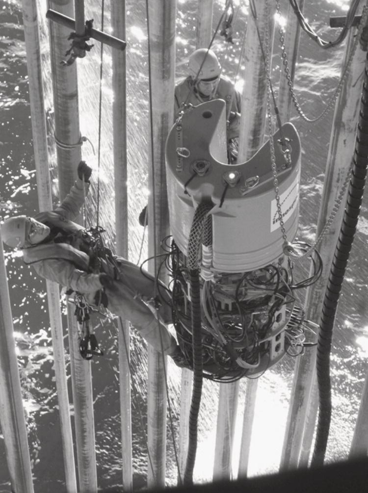 Two individuals are suspended in a marine environment, engaged in a specialized operation. They are wearing protective suits and helmets, emphasizing safety in a challenging working condition. Attached to a harness, one person is being carefully lowered with a piece of equipment resembling a large, cylindrical device, which features multiple cables and electronic components. Surrounding them are tall structures, possibly part of an offshore rig, indicating the setting’s industrial nature. Reflections on the water surface add a dynamic element to the scene.