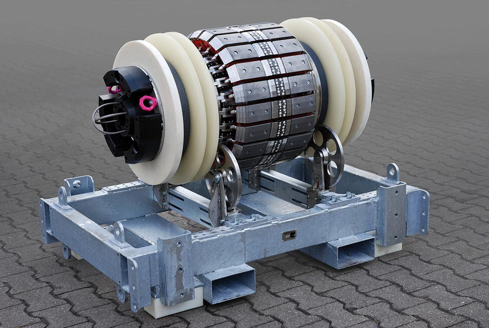 An object features a cylindrical core surrounded by several circular bands, which appear to be integral components of a mechanical system. Two large, smooth wheels flank the core, possibly enabling movement or rotation. The entire assembly rests on a rectangular metallic frame, with various attachment points and a sturdy design, suggesting it is designed for support or transport. The ground surface appears textured, indicating an environment suitable for industrial or engineering applications. The overall structure hints at advanced engineering with specific functional purposes.