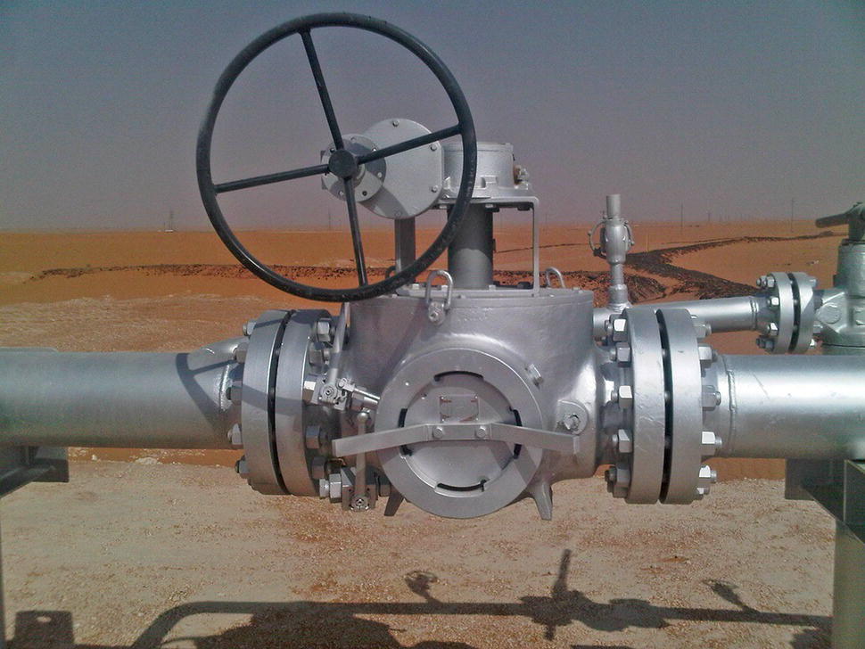 A large industrial valve is prominently featured, equipped with a circular wheel handle for manual operation. The valve body is metallic, with a smooth, shiny surface, indicating its robust construction. Two flanged pipe connections extend from either side of the valve, designed to facilitate the flow of liquids or gases. In the background, a flat landscape is visible, possibly a coastal area with a clear sky and distant lines that suggest a separation between land and water. The overall setup reflects a functional, industrial environment.