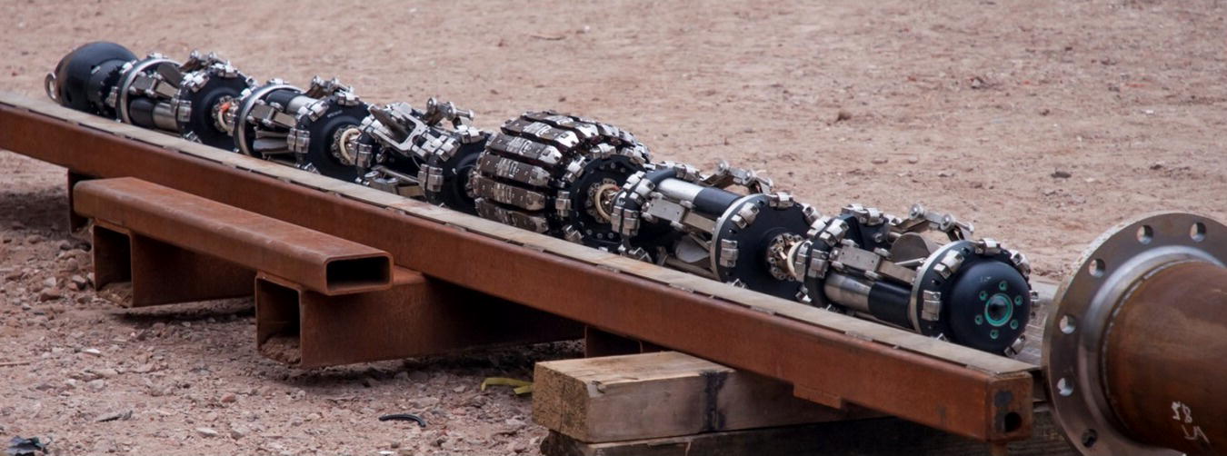 A long cylindrical object rests horizontally on a platform, supported by blocks. The object is segmented with multiple sections, each containing intricate mechanical components, including gears and chains that interconnect them. These components suggest functionality related to machinery or engineering. The background appears rugged and unrefined, enhancing the industrial feel. Each segment has a distinct arrangement of parts, indicating a complex design likely intended for a specific mechanical purpose or advancement in technology.