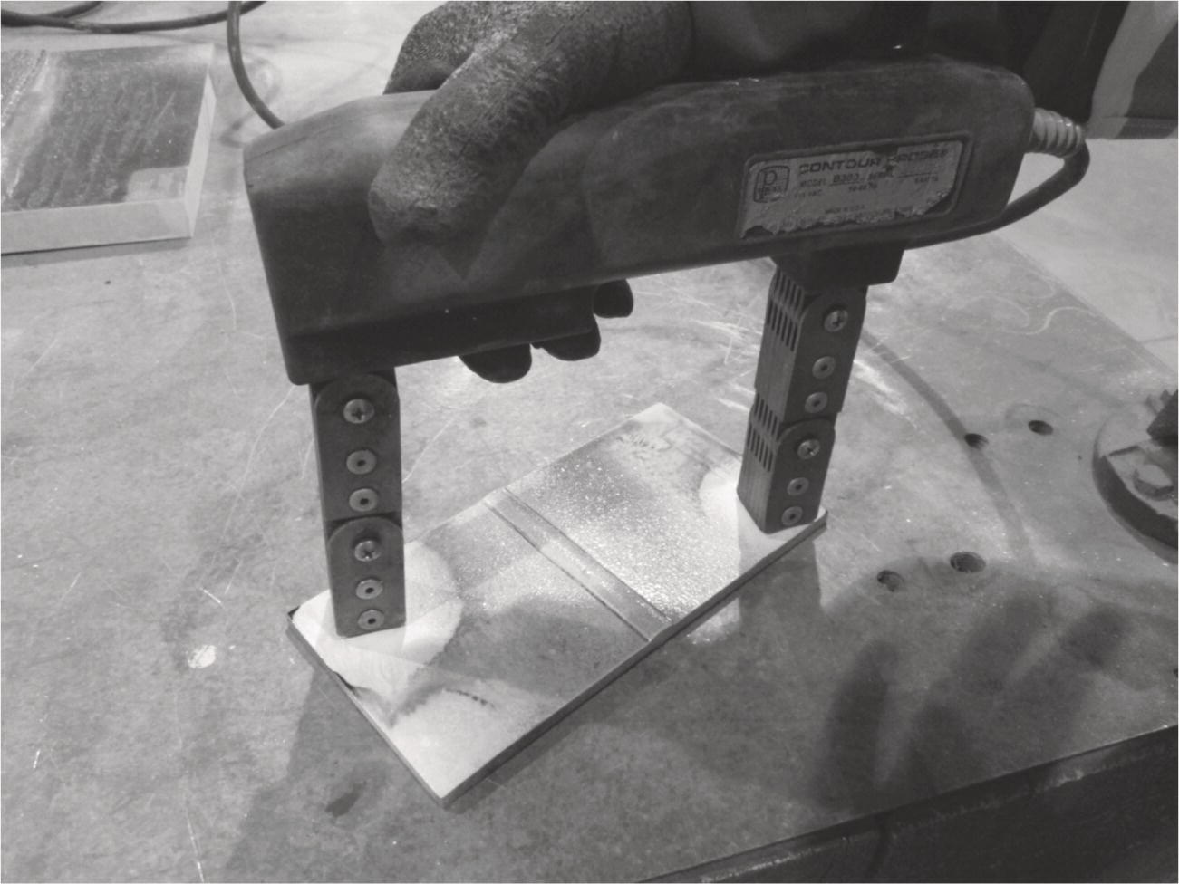 A hand grips a device designed for welding or joining materials, featuring two parallel metal arms with round ends. The device is positioned over a workpiece, which is a rectangular metal plate resting on a surface. The plate exhibits a shiny, smooth line across its centre, suggesting recently applied heat or treatment. The background reveals a workshop environment, with tools and machinery visible, reflecting an industrial setting. The glove on the hand indicates a focus on safety during operation.