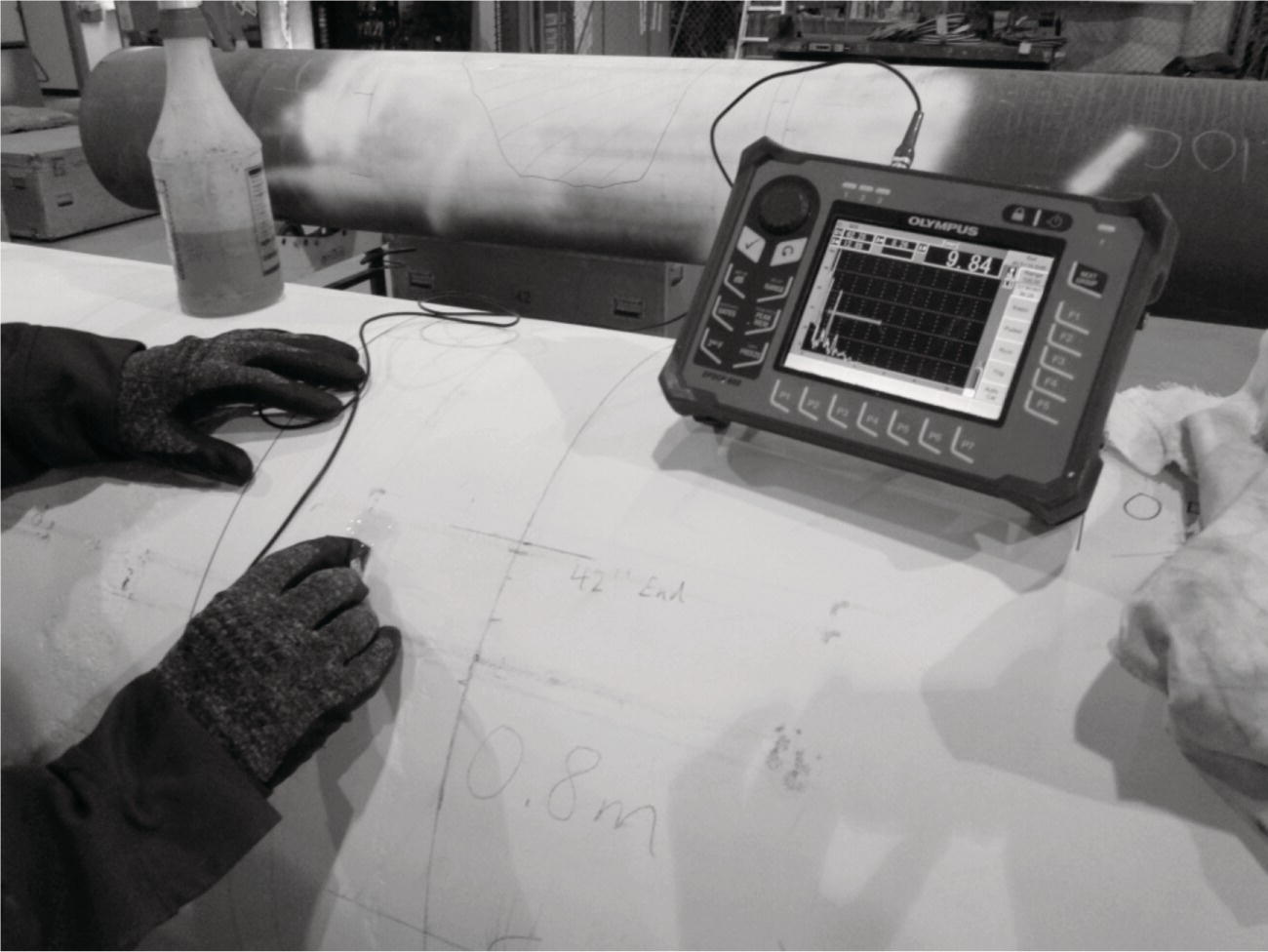A workshop environment has a person wearing gloves interacting with a handheld device placed on a cylindrical surface. The device, likely an ultrasonic testing instrument from Olympus, displays a graphical readout that appears to indicate measurements. Nearby, a container is partially visible, suggesting a preparation for testing. Handwritten markings on the cylindrical surface provide reference points, including 42 inch End and a measurement of 0.8 m, indicating potential inspection zones or features of interest. The focus on precise measurement suggests a critical assessment of material integrity.