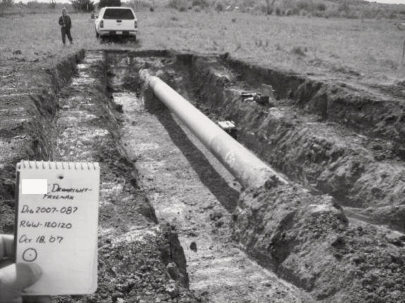 A trench has been excavated, revealing a long, metallic pipe running horizontally through the ground. The trench is surrounded by disturbed earth, indicating recent digging. In the foreground, a hand holds a notepad with handwritten notes and a date, October 18, 2007, along with additional annotations. In the background, a person stands near a vehicle parked on the open landscape, which features grass and a clear sky. The setting appears to be rural, with minimal vegetation and a wide expanse of land visible.