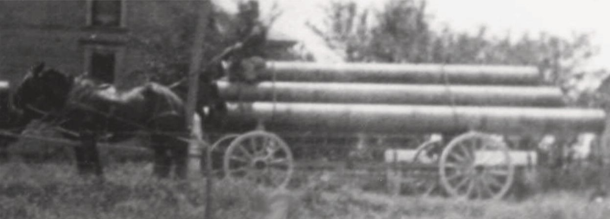 A horse-drawn wagon carries large cylindrical objects piled on a wooden cart. The horse, equipped with a harness, stands beside the wagon, which has two large wheels. In the background, an indistinct building is visible, surrounded by trees and shrubbery. The setting appears rural, with a grassy area surrounding the wagon, creating a sense of a bygone era.