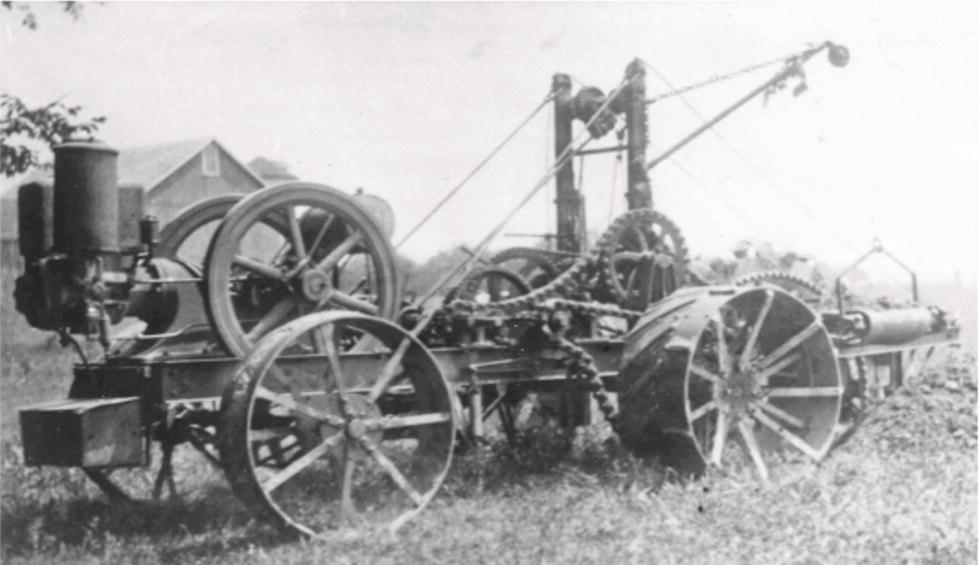 A large, vintage agricultural machine stands on a grassy field, featuring a series of large wheels that are spoked and robust. The machine has a prominent engine on the left side, with a cylindrical component contributing to its mechanical design. Chains and gears are visible, indicating a complex mechanism for farm work. Above, there are various levers and pulleys, suggesting the capability for multiple functions. In the background, a farmhouse can be seen, hinting at the rural setting of this historical farming implement.