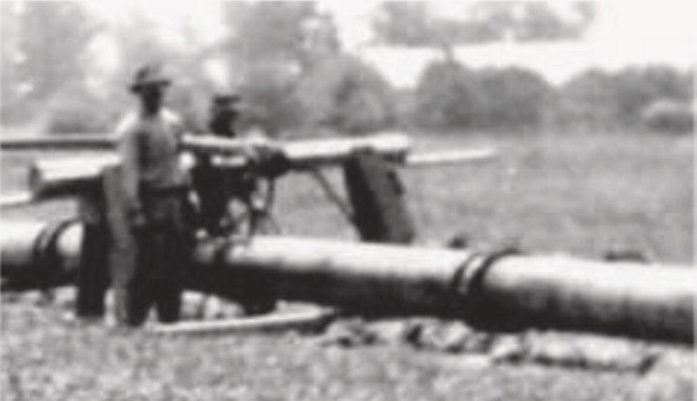 Two men, one leaning casually against a large cannon-like structure, stand beside a long, heavy barrel resting on the ground. The first man, wearing a wide-brimmed hat, gazes towards the distance. The second man, positioned slightly behind, operates a mechanism mounted on the barrel. The background features a blurred landscape, suggesting an outdoor setting with trees and possibly buildings in the distance, creating a historical atmosphere reminiscent of earlier military technology or industrial endeavours.
