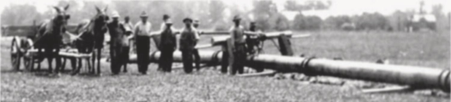 A group of men stands beside a large piece of machinery in a rural setting. They are dressed in early 20th-century work attire, including hats and overalls. In the background, a horse-drawn cart rests, with a horse harnessed to it. The landscape features tall grass and trees in the distance, suggesting a farmland environment. The equipment, resembling a steam engine or an industrial apparatus, stretches along the ground, indicating a scene focused on labour and machinery from that era.