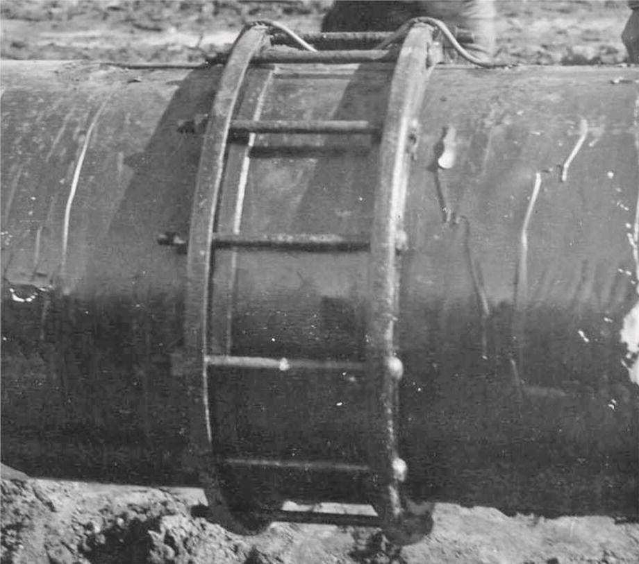 A large cylindrical pipe rests on the ground, featuring a robust metal band secured around its midsection. The band is equipped with multiple horizontal rungs for climbing or support. The surface of the pipe appears worn and glossy, indicating exposure to the elements. Soil and earth are visible around the base, suggesting it is partially buried or recently installed. In the background, another person is present, adding context to the scene, which appears industrial or construction-related.