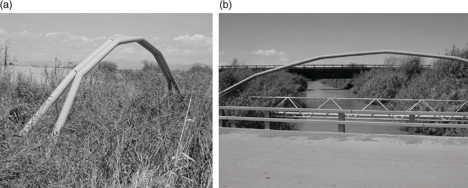 An image presents two photographs labelled a and b. In a, a curved structure made of metal pipes stands amidst tall grass and vegetation under a cloudy sky, leaning slightly to one side. In b, the same curved metal structure is visible again, this time situated over a body of water, with additional metal framing in the foreground, supported by pillars. The backdrop features a bridge that spans the scene above the waterway, surrounded by trees and sparse vegetation. The contrast highlights the environment around the structures.
