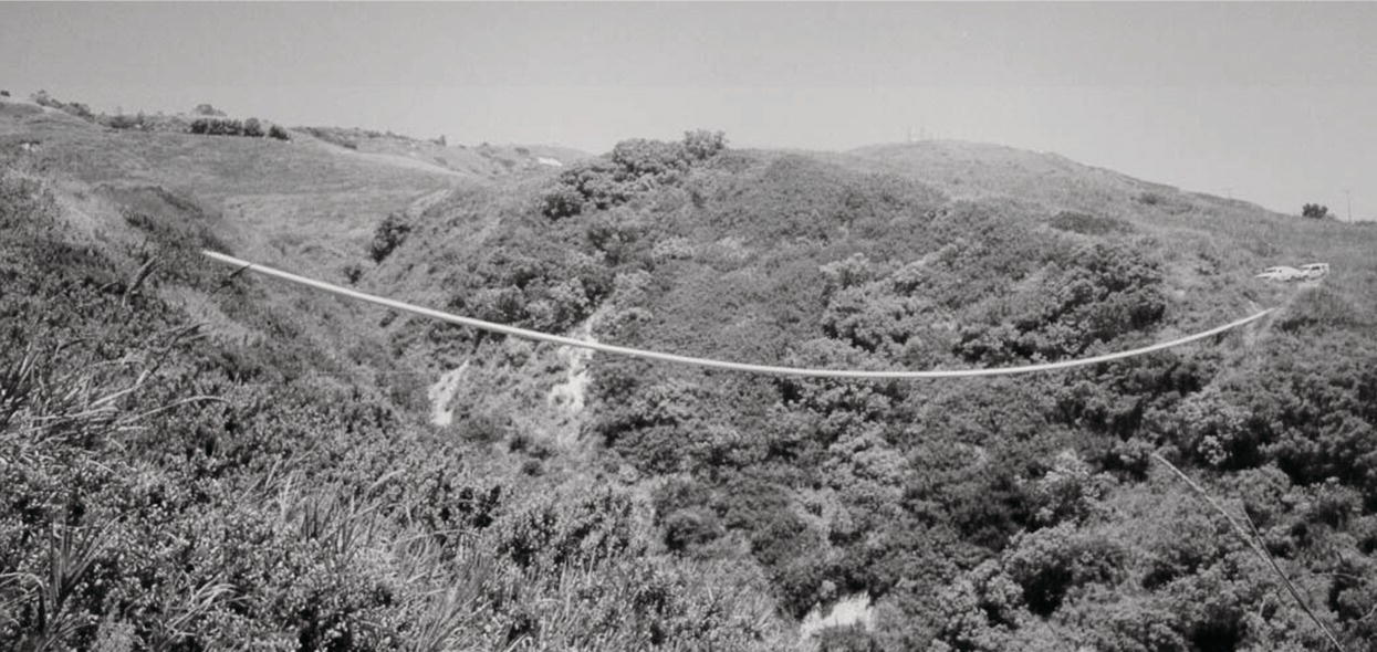 A photograph captures a landscape characterized by rolling hills covered in dense foliage. A long, curved cable stretches across a deep valley, connecting two elevated points. The surrounding terrain features patches of greenery and light soil, with a dirt path winding up one side of the hillside. The overall scene conveys a sense of adventure and engineering, emphasizing the contrast between the natural environment and the human-made structure. The curvature of the cable adds a dynamic aspect to the composition.