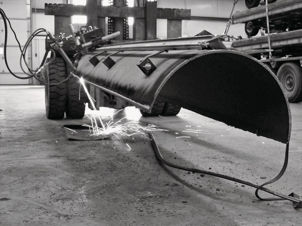 A large, curved metal structure sits on the floor of a workshop, supported by heavy machinery on the left. The structure is mounted on wheels, emphasizing its substantial size. Sparks fly from the lower part where welding tools are in use, illuminating the surroundings with bright flashes. Various tools and equipment are arranged around the area, indicating an active work environment. The background features stacked machinery, contributing to a sense of industry and manufacturing. The atmosphere captures an essence of craftsmanship and heavy-duty work.