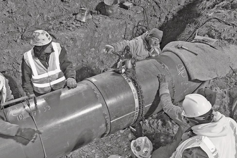 A group of workers is engaged in a construction project involving a large pipe. The scene is set in a trench, with several individuals wearing safety helmets and reflective vests. One worker appears to be operating a mechanical tool attached to the pipe, while others assist in handling it. The pipe has markings indicating measurements or specifications. The trench walls are rough, suggesting substantial excavation effort. Tools and equipment are scattered around, emphasizing the construction environment and teamwork essential for the task.