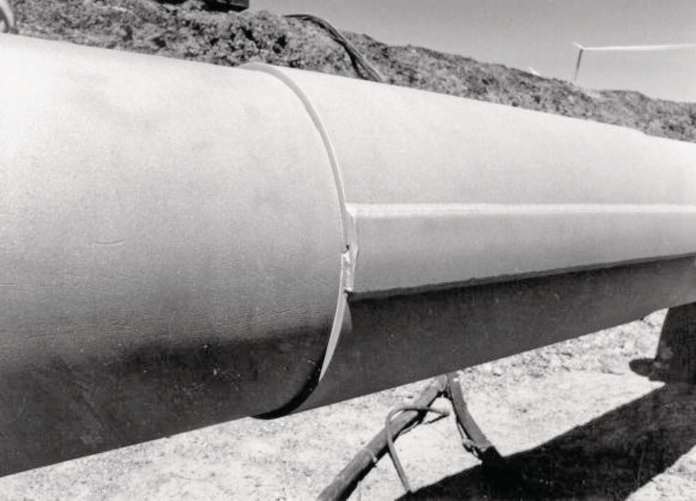 A large cylindrical pipe is featured prominently, resting on a flat, dry surface. There is a noticeable vertical crack or seam along its length, indicating potential damage or weakness. A thin band or strap encircles the pipe at the cracked area, suggesting an attempt at reinforcement or repair. The background consists of rough terrain, likely soil or gravel, with some equipment partially visible. Bright sunlight casts distinct shadows, enhancing the texture and surface details of the pipe and surrounding ground.