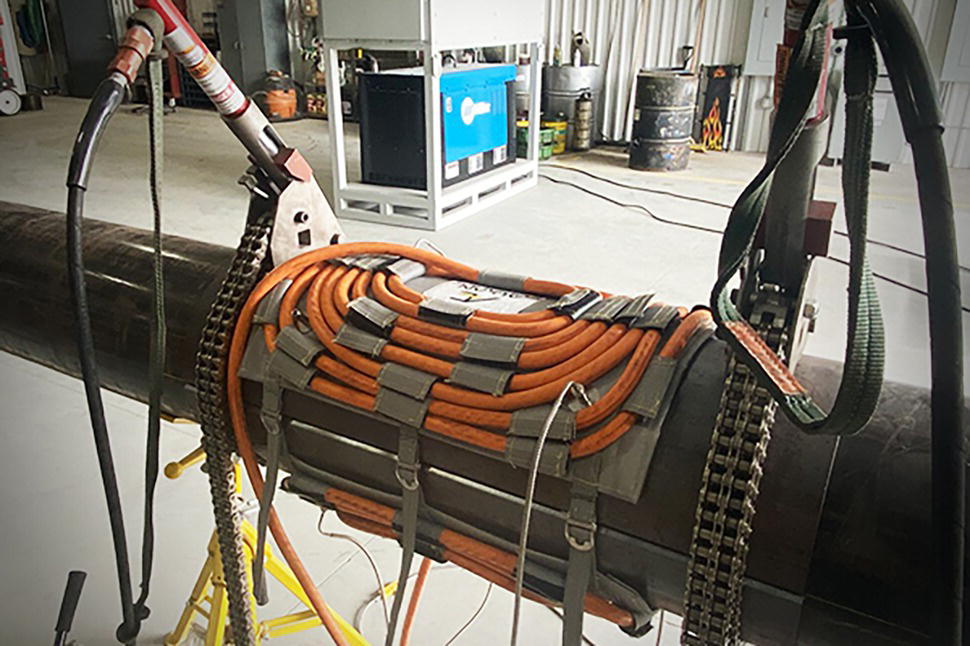 A large cylindrical object lies horizontally in a spacious workshop filled with various tools and equipment. On top of the cylinder, a dark, rectangular device is carefully secured, featuring several coiled hoses arranged neatly in a circular pattern. Flexible cables extend from the device, connected to various points, suggesting a complex mechanical setup. Behind, barrels are lined up, hinting at industrial use. The background presents a faint outline of equipment, indicating a busy work environment focused on technical operations.