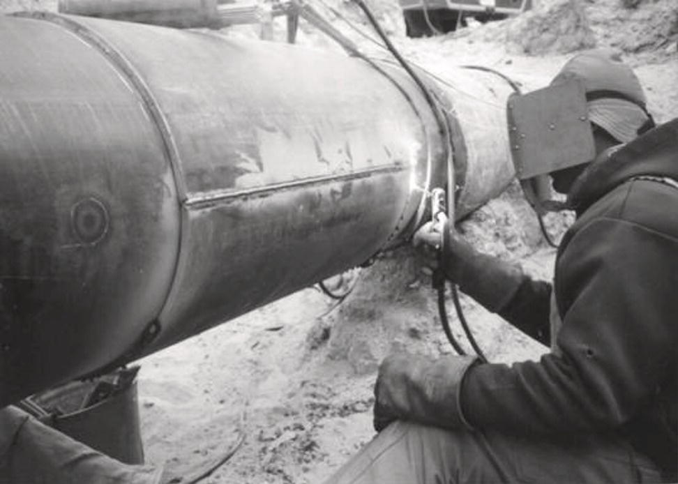 A worker engages in the process of welding a large, cylindrical pipe. Clad in protective gear, including a helmet and gloves, the individual focuses intently on the task at hand. Sparks fly as the welding torch makes contact with the metal surface, illuminating the area around the joint. The background reveals a sandy environment, suggesting work in an outdoor setting. Tools and hoses are arranged nearby, indicating the complexity of the operation and the need for specialized equipment in this industrial activity.