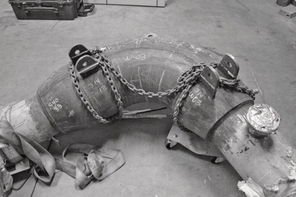 An image presents a photograph of a large curved pipe fitting or elbow joint lying on a concrete floor. The fitting has heavy metal chains wrapped around both ends for handling or transport. The pipe section appears industrial-grade with visible flanges at each end and what seems to be a small circular access port or gauge mount on the right side. The surface of the pipe has weathered markings and some apparent damage or wear. Various tools or metal pieces lie scattered around the pipe on the floor, suggesting an industrial or construction setting.