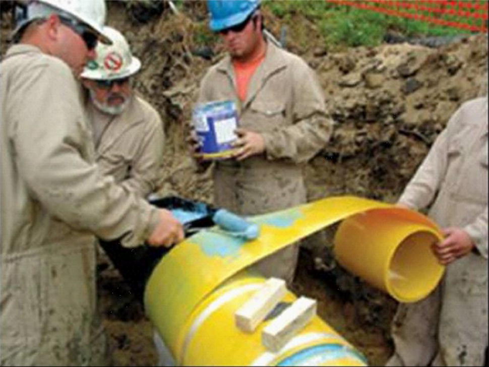 A photograph captures construction workers in hard hats and work clothes installing a large pipe section in an excavated trench. Three workers are visible, one on the left manipulating something on the pipe, another in the centre holding a measurement equipment or documentation, and a third partially visible on the right handling a curved pipe component. The scene appears to be part of a utility installation project, with disturbed soil visible in the background. The workers are focused on properly positioning or connecting the cylindrical pipe segments.