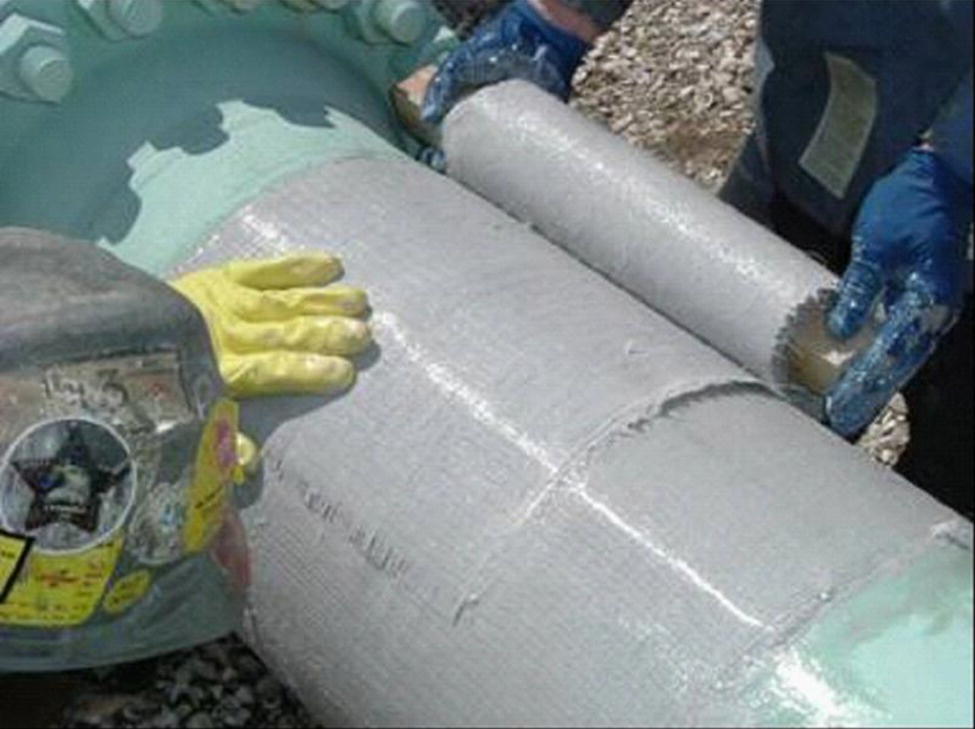 A photograph captures a close-up of construction work on a large pipe. A worker’s gloved hand is visible on the left side touching the pipe’s surface. The cylindrical pipe appears to have some markings or text imprinted on it. There’s evidence of joint preparation or sealing work happening along the pipe’s length, with what seems to be a fresh application of coating or sealant material visible. Construction materials or equipment can be partially seen in the background. The image documents industrial pipeline installation or maintenance procedures.