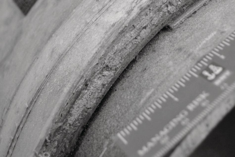 An image presents an extreme close-up of a pipe or cylindrical object’s edge. A curved surface with visible texture and a seam or joint runs across the frame. In the lower right corner, a measuring device with metric markings is positioned against the surface, likely for measuring dimensions or defects. The surface has noticeable textural variations along the curved edge, suggesting possible corrosion, damage, or material transition. The photograph seems to document an inspection or assessment of the structural condition of this industrial component.
