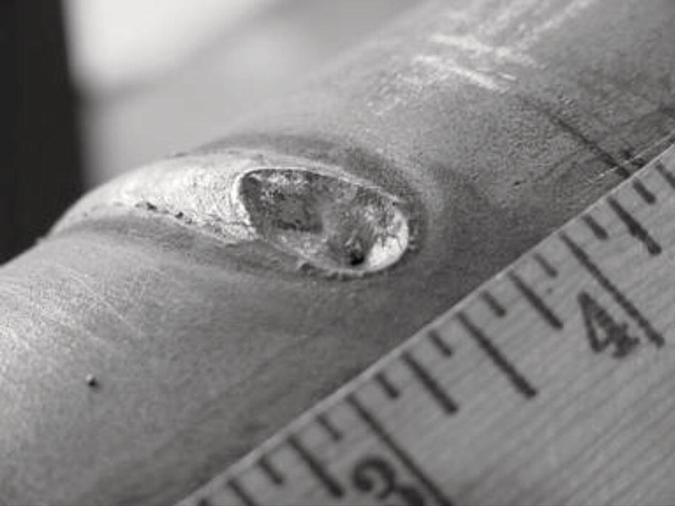 A photograph captures a close-up of a metal pipe or cylinder with a noticeable defect or cavity on its surface. The oval-shaped depression appears to be damage, possibly corrosion or a manufacturing flaw. A measuring ruler is positioned along the bottom edge of the frame, with visible measurement markings from 2 to 4 inches, providing scale reference for the defect. The metallic surface has a slightly textured appearance with some visible scratches or markings. This appears to be documentation of material damage for inspection or quality control purposes.