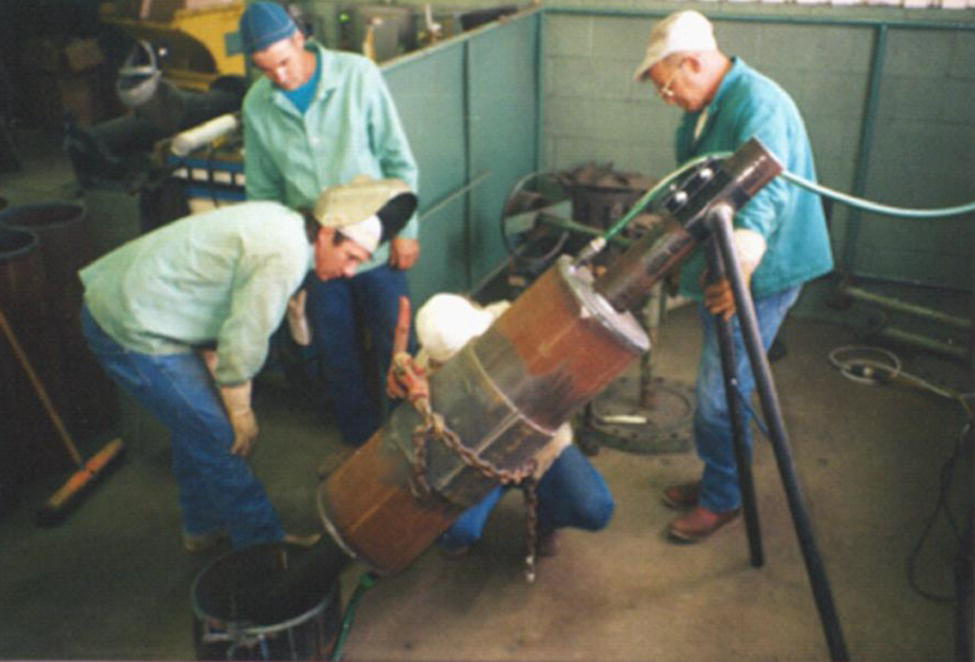 A photograph captures three workers in an industrial setting involved in a metalworking or foundry operation. They wear protective clothing and caps while handling a crucible or large metal container that’s pouring or transferring molten material into a receptacle below. The workers are positioned around the equipment, with one operating a mechanism with a hose or cable attached. The environment looks confined with walls visible in the background, typical of a manufacturing or metallurgical facility. This appears to document a metal casting or smelting process requiring careful coordination among the workers.