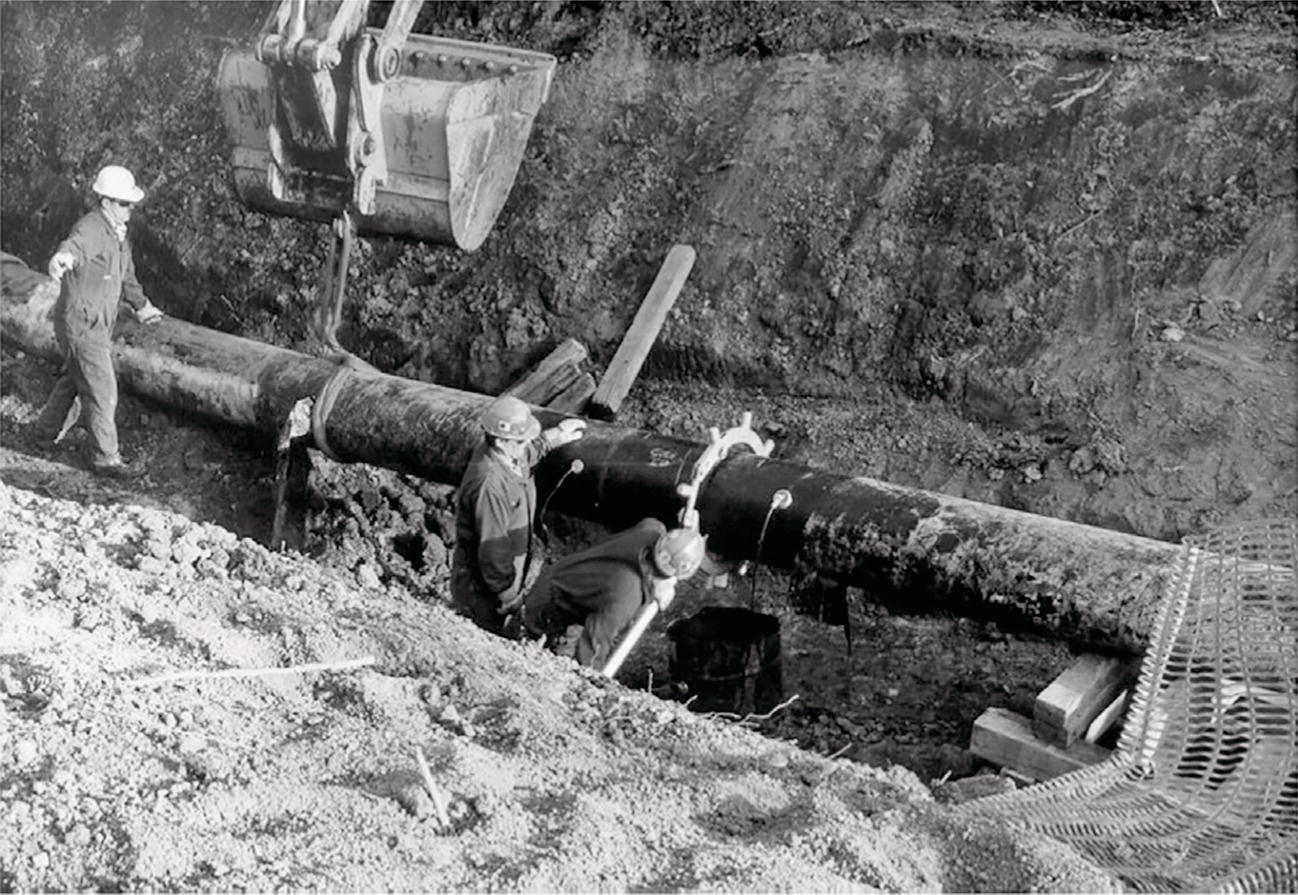 A photograph captures pipeline construction workers in a trench. Two workers in hard hats and work clothes are positioned around a large diameter pipe being installed underground. An excavator bucket is visible at the top of the frame. The workers appear to be aligning or connecting pipe sections within the excavated ditch. Soil and debris are piled along the edge of the trench, and what looks like safety mesh or grating is visible at the bottom right corner of the excavation site.