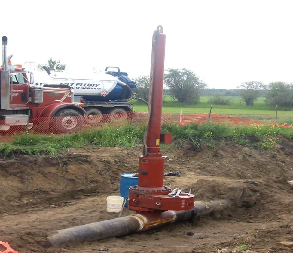 A photograph captures pipeline maintenance equipment in a rural setting. A vertical pipe tapping or hot tap machine is connected to a horizontal underground pipeline in an excavated trench. The equipment appears to be drilling into or working on the pipeline while it remains in service. In the background, a large truck with Fleury visible on its side is parked near the work site. The surrounding area consists of open countryside with some trees visible on the horizon and a fence line running parallel to the work area.