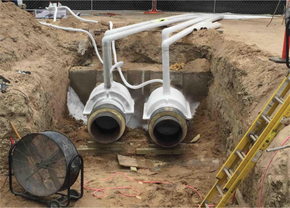 A photograph features an excavated trench containing two large parallel pipes with attached valves or fittings. The pipes appear to be part of a utility installation, possibly for water, steam, or district heating systems. The trench has been dug with vertical walls exposing the soil layers. A portable fan or blower sits on the left side of the trench floor, likely for ventilation purposes. A ladder is visible on the right edge, providing access to the excavation. Small-diameter pipes or conduits extend from the main pipes, creating a branching configuration.