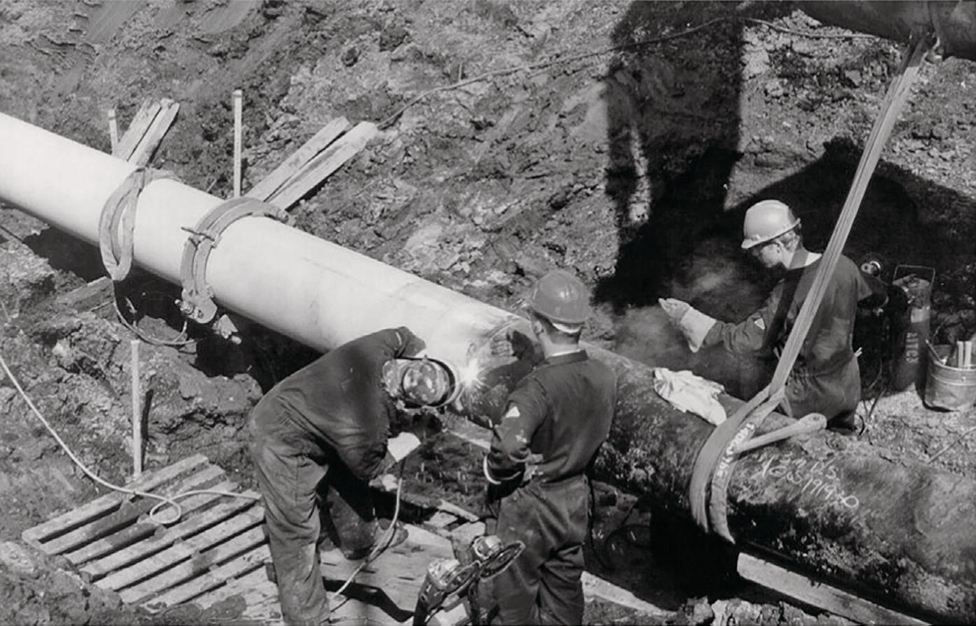 A photograph captures workers in hard hats performing pipeline construction or maintenance in an excavated trench. Three workers are visible, with two in the foreground working directly on a connection point between pipes. One appears to be using tools at the joint where a clean, light-coloured pipe meets an older, rough-textured pipe. A wooden platform or grating lies on the trench floor providing a stable working surface. The trench walls are clearly visible, exposing layers of soil. The workers are wearing protective gear as they complete a pipe installation or repair operation.