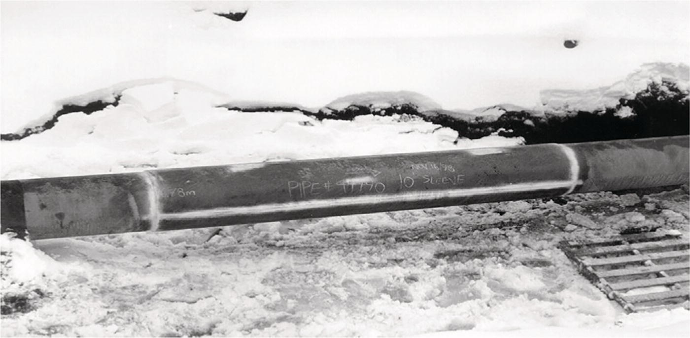 A photograph captures a pipeline partially covered with snow. The exposed section of pipe has markings or text written on its surface, though the specific text is difficult to fully read. The pipe is laid in what looks like an excavated trench with rocky, disturbed soil visible below. At the bottom right corner, there appears to be a small section of grating or ladder. The contrast between the dark pipe, light snow cover, and textured ground creates clear definition in the image.