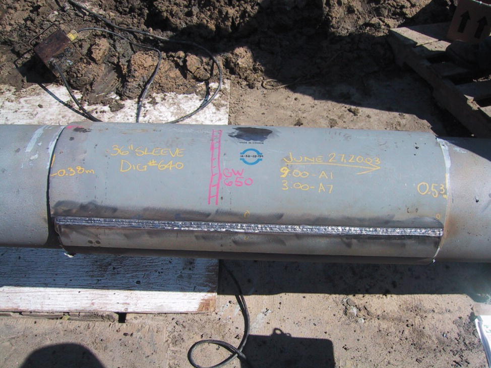 A photograph features a section of large diameter pipeline in an excavated trench. The pipe has handwritten markings or measurements written directly on its surface. A welded seam runs horizontally along the pipe, likely a repair or connection point. The surrounding environment includes exposed soil walls of the trench. Some cables or wires are visible near the bottom of the image, possibly related to welding or inspection equipment. A wooden board or plank appears to be positioned beneath a portion of the pipe, potentially for support during the installation or repair process.