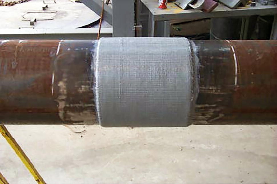 An image captures a pipe or cylindrical component with a repair or reinforcement section in the middle. A light-coloured patch or wrap material has been applied around the central portion of the dark pipe, creating a visible repair joint. The pipe appears to be positioned horizontally in an industrial or workshop setting. The repair section has a textured surface, suggesting it might be fiberglass, composite material, or specialized tape used for pipe repairs. The surrounding environment includes work surfaces or equipment, though they're partially visible in the background.