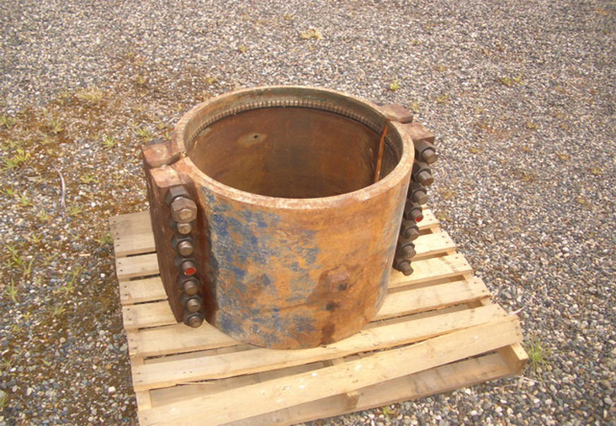 A photograph of a large cylindrical pipe coupling or repair sleeve resting on a wooden pallet. The metal sleeve has a series of bolts along one side that is used for tightening or securing it around a pipe. The coupling has a circular opening and seems designed to join or repair sections of pipeline. The pallet sits on a gravel or aggregate surface. The coupling appears weathered or used, with a rough texture on its exterior surface.